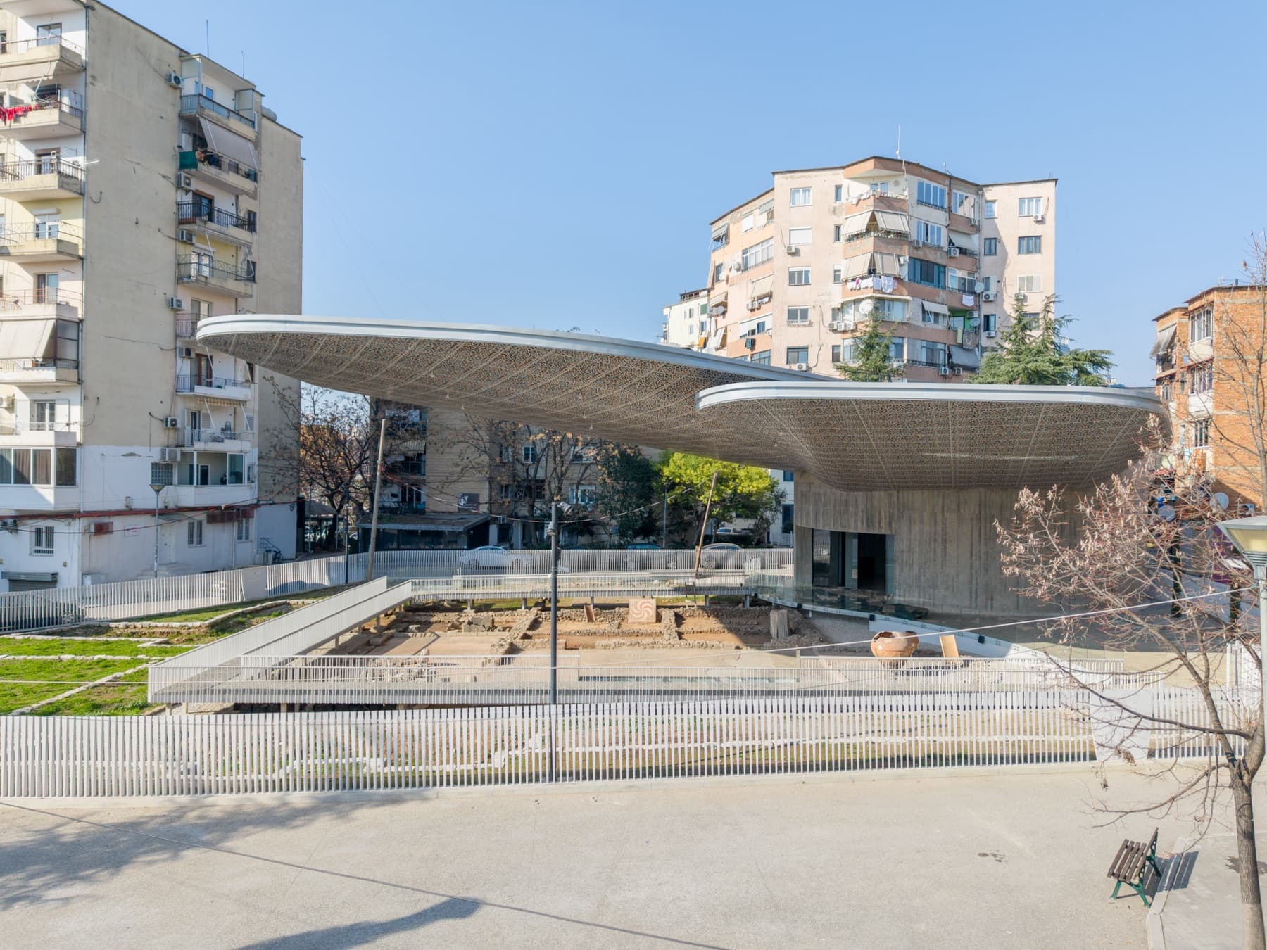 Pamje e jashtme e pavijonit të Mozaikut të Tiranës, me tendën e tij të perforuar që rri pezull mbi sitin arkeologjik, të kornizuar nga pallatet e Tiranës në një ditë të kthjellët.