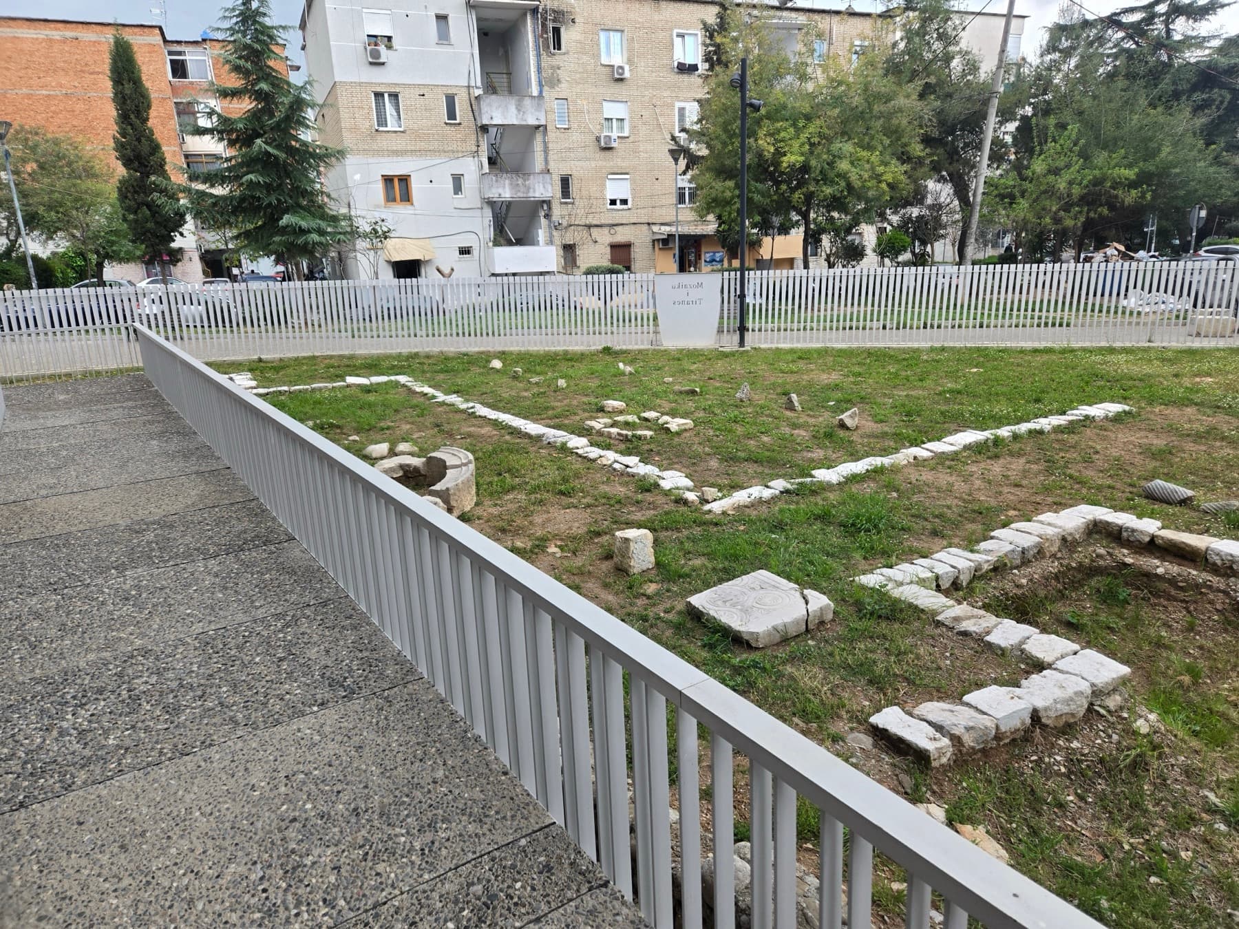 Stone foundation remains of the Roman villa laid out in grass, bordered by the protective fence, with Tirana residential blocks behind.
