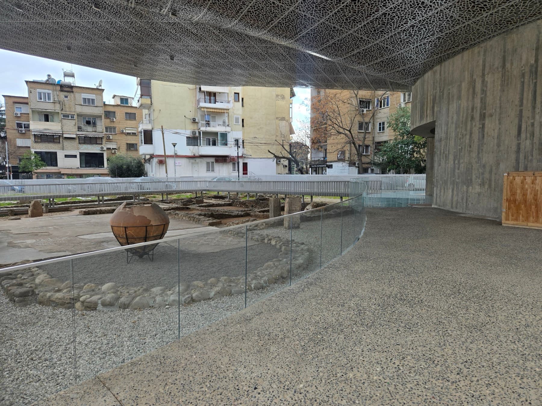 Interior of the protective pavilion with an amphora at centre and glass railings around the excavation, evoking the visitor tour.
