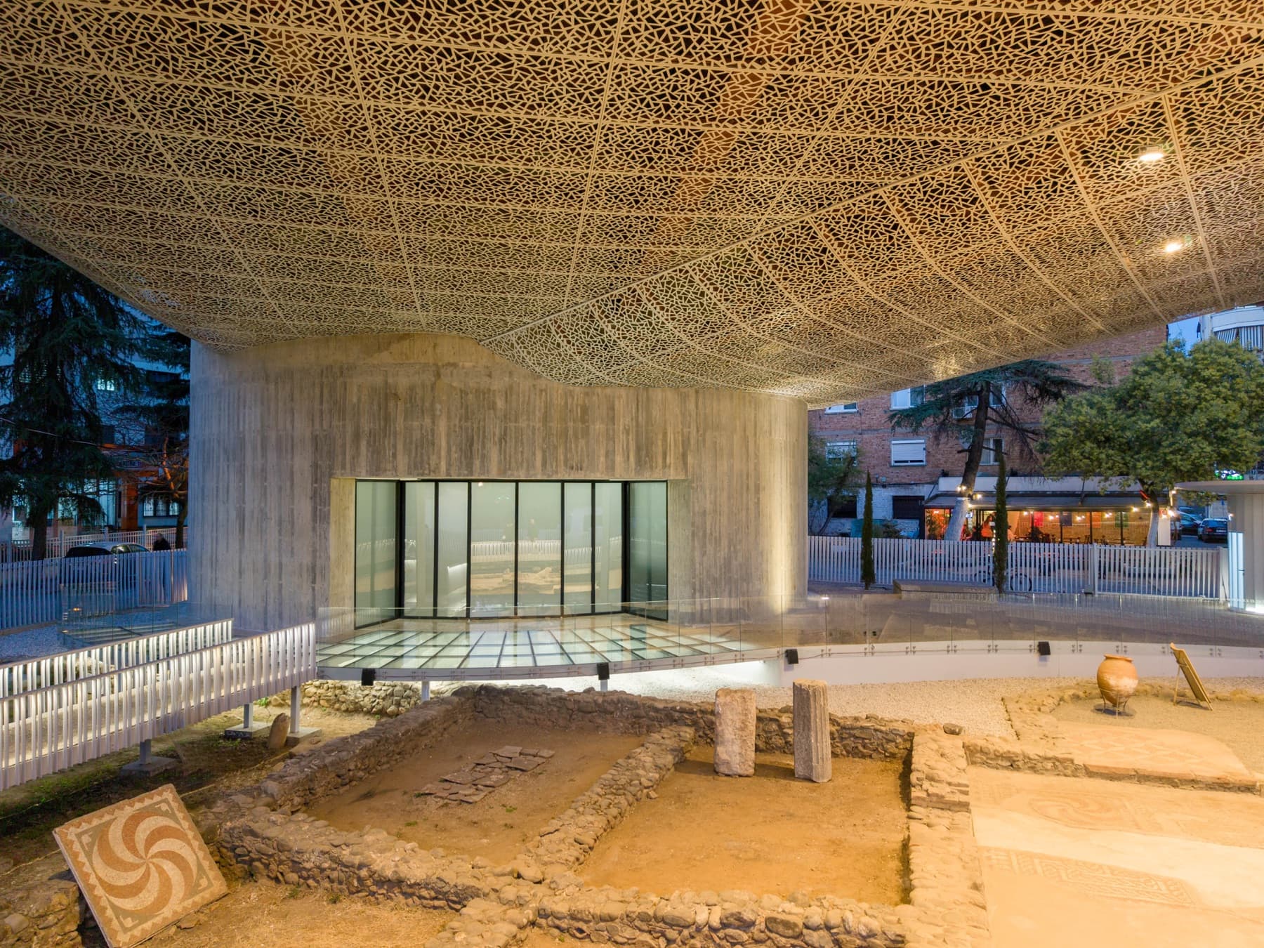Evening view inside the pavilion: the illuminated concrete gallery volume and glass-floored walkway above the Roman excavation, with a mosaic panel and amphora on display.