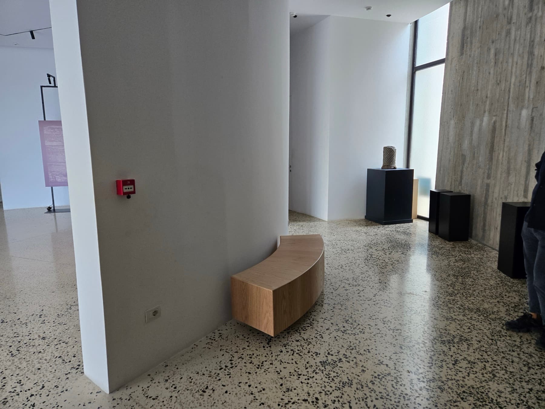 Bright museum interior with terrazzo flooring, white walls, a curved wooden bench, and stone artefacts on pedestals.