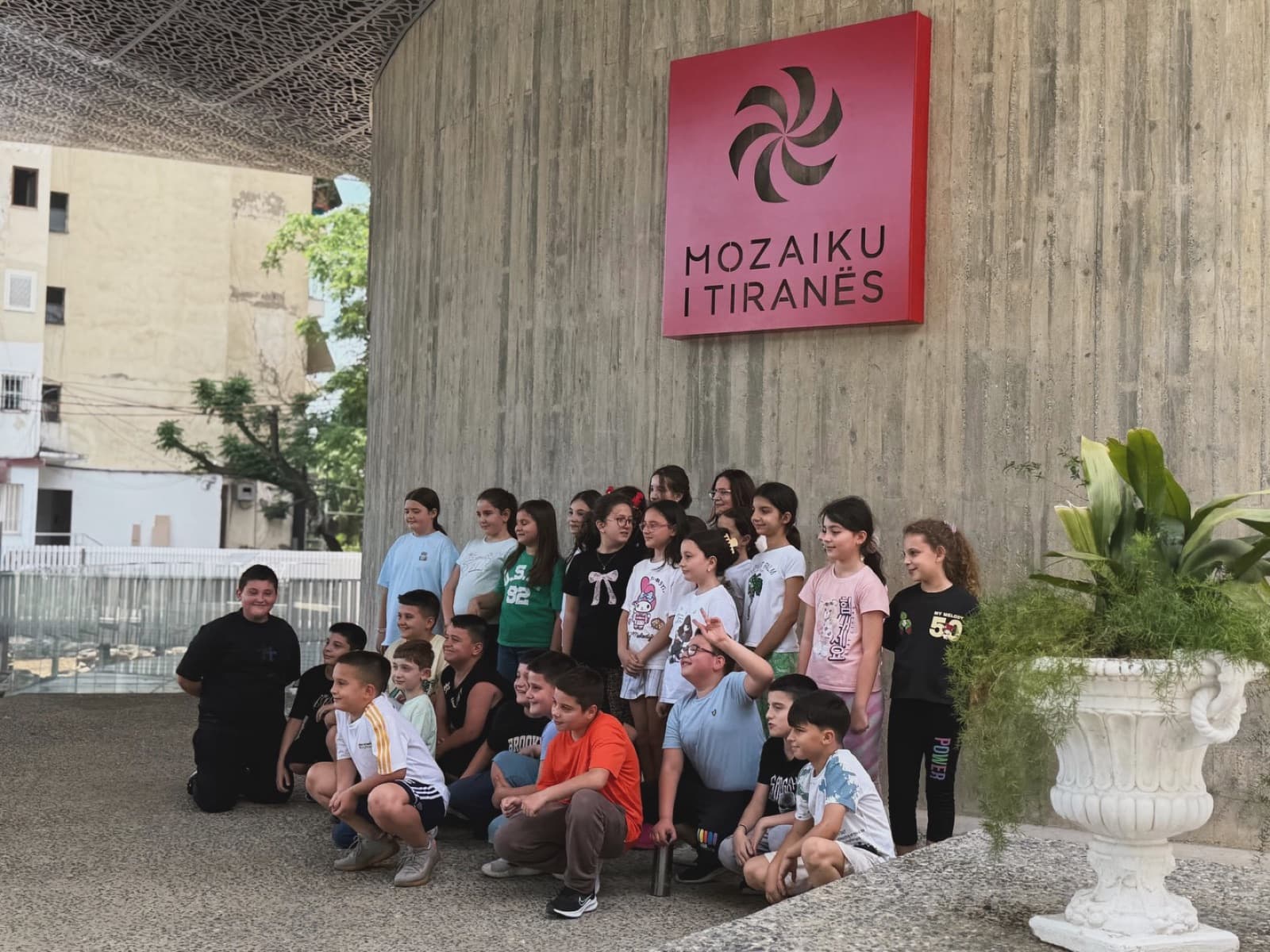 Schoolchildren from Pjetër Budi School gathered beneath the Mozaiku i Tiranës sign during a Little Archaeologist visit.