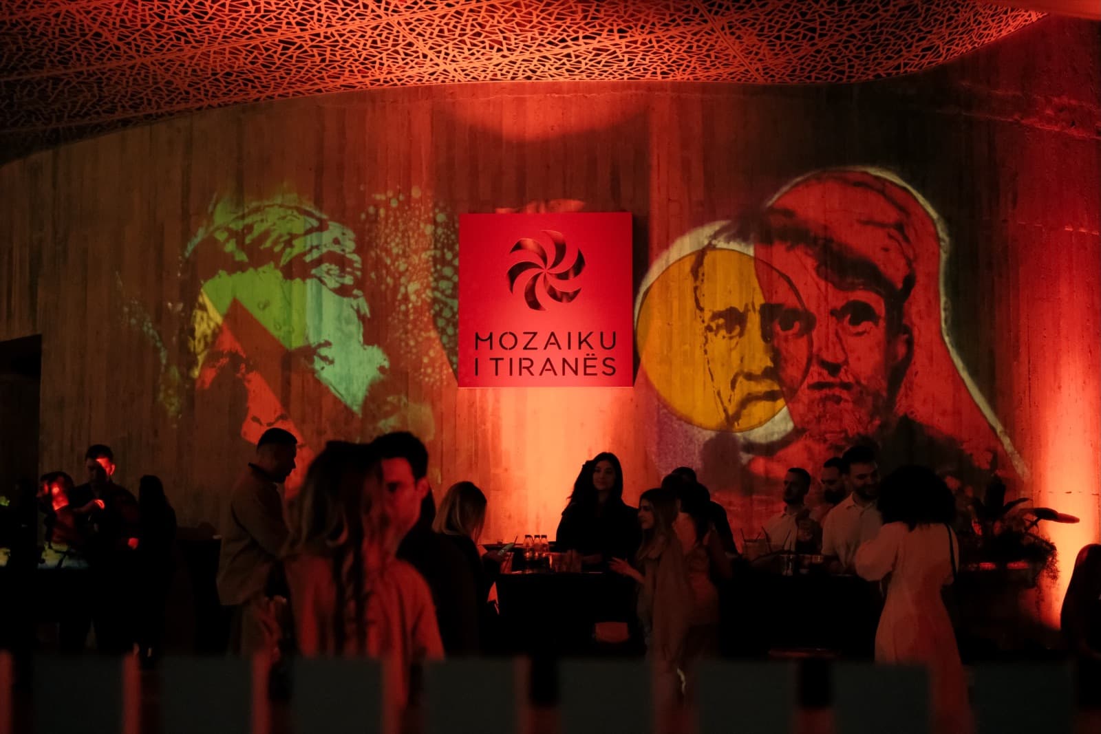 Evening crowd at the Museum Week 2026 ‘Beautiful Minds of Albania’ event, with portraits projected onto the pavilion wall beside the illuminated Mozaiku i Tiranës sign.
