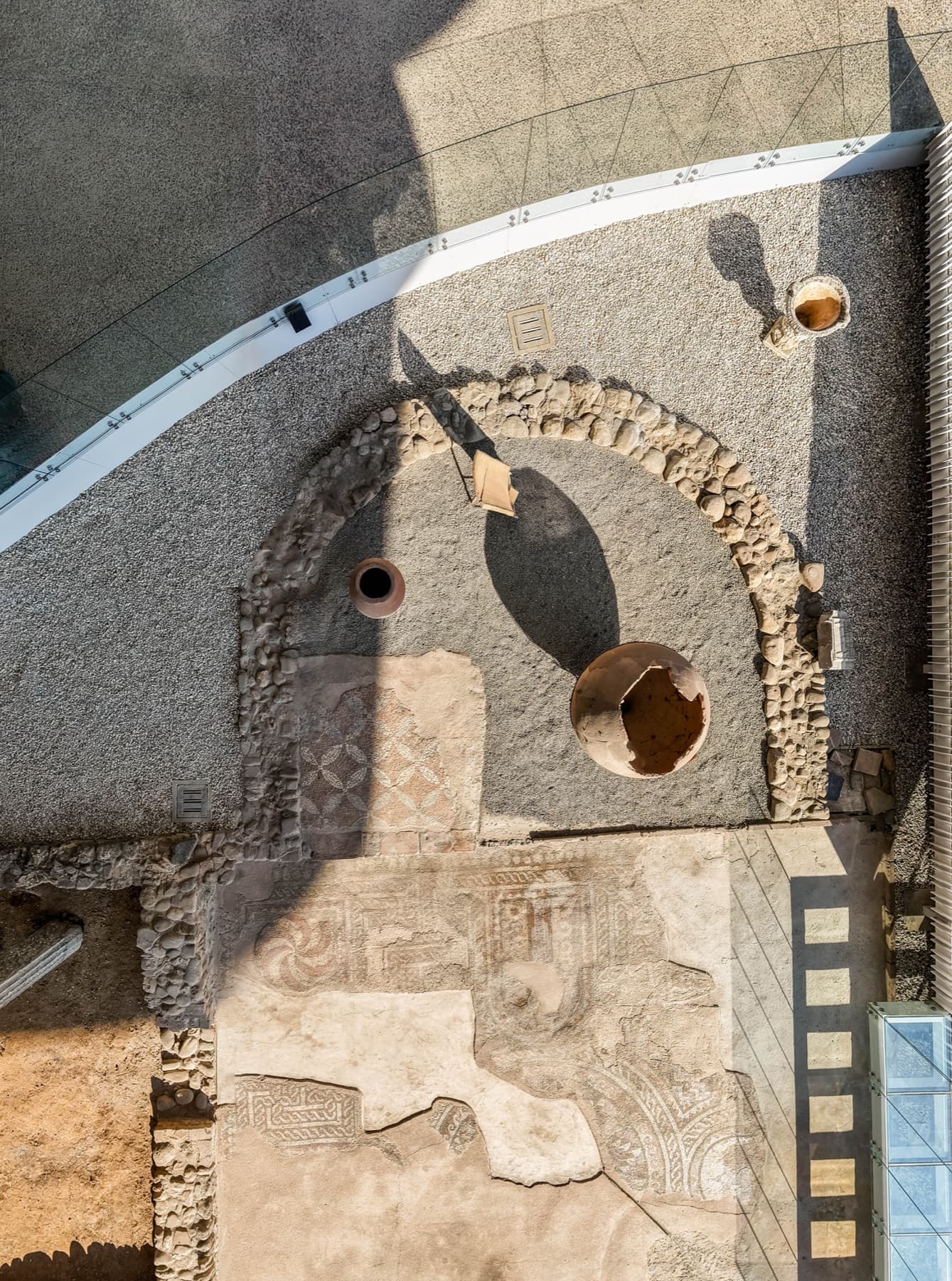 Overhead view of the excavation showing the Roman villa's stone foundations, patterned mosaic floors and amphorae, framed by the glass-railed visitor walkway.