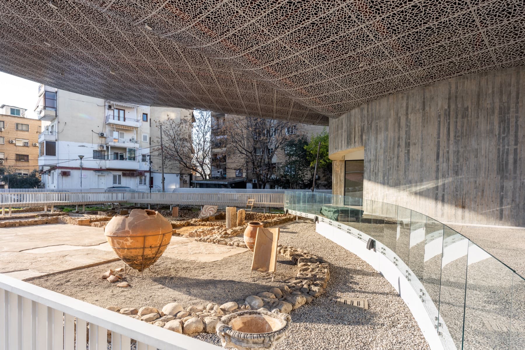 The Roman villa excavation beneath the pavilion's perforated canopy, with large terracotta storage jars set among the stone foundations along the glass-railed walkway, apartment blocks beyond.
